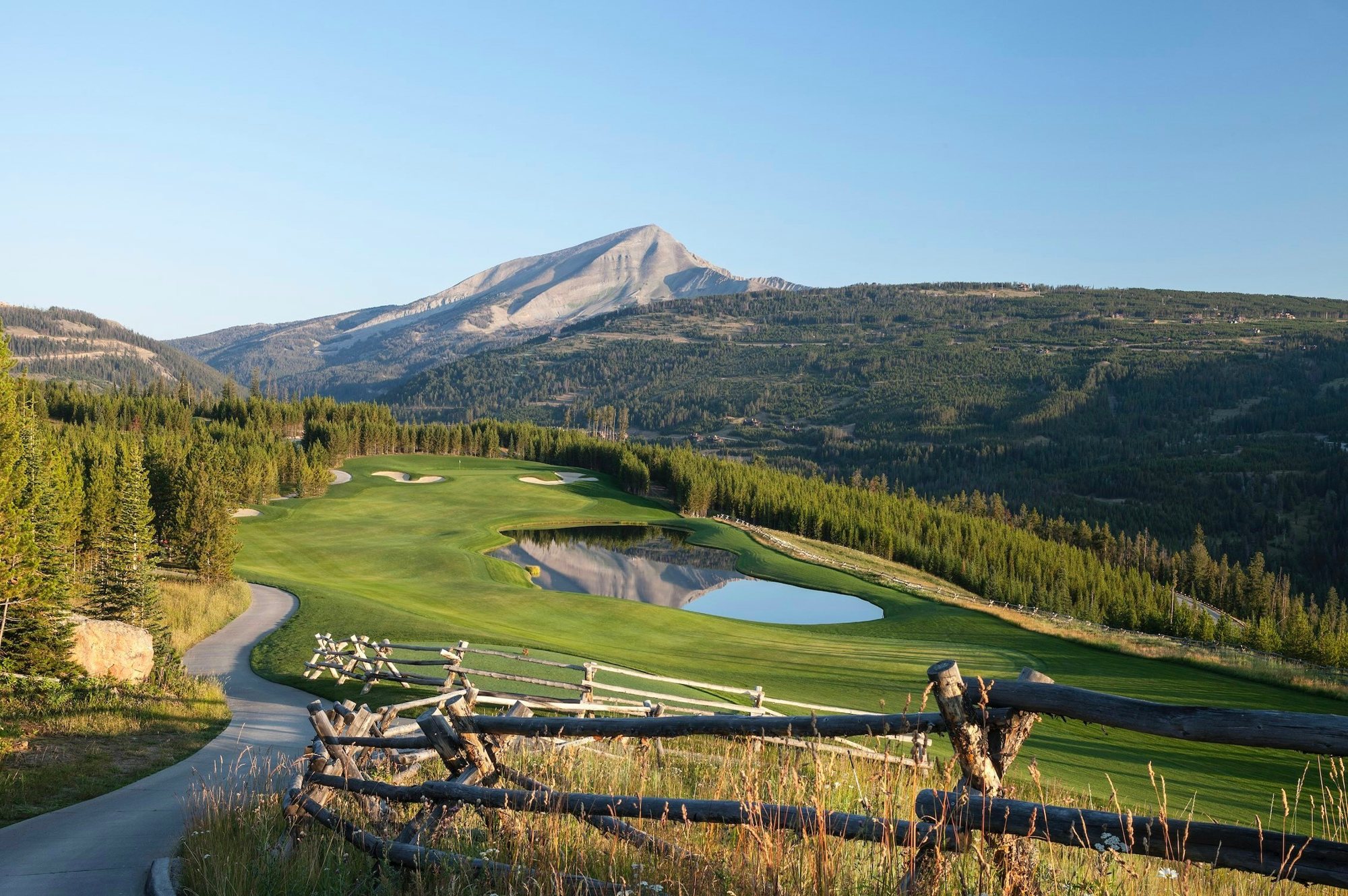 A Yellowstone Club golf fairway and pond below Lone Peak, framed by a split-rail fence