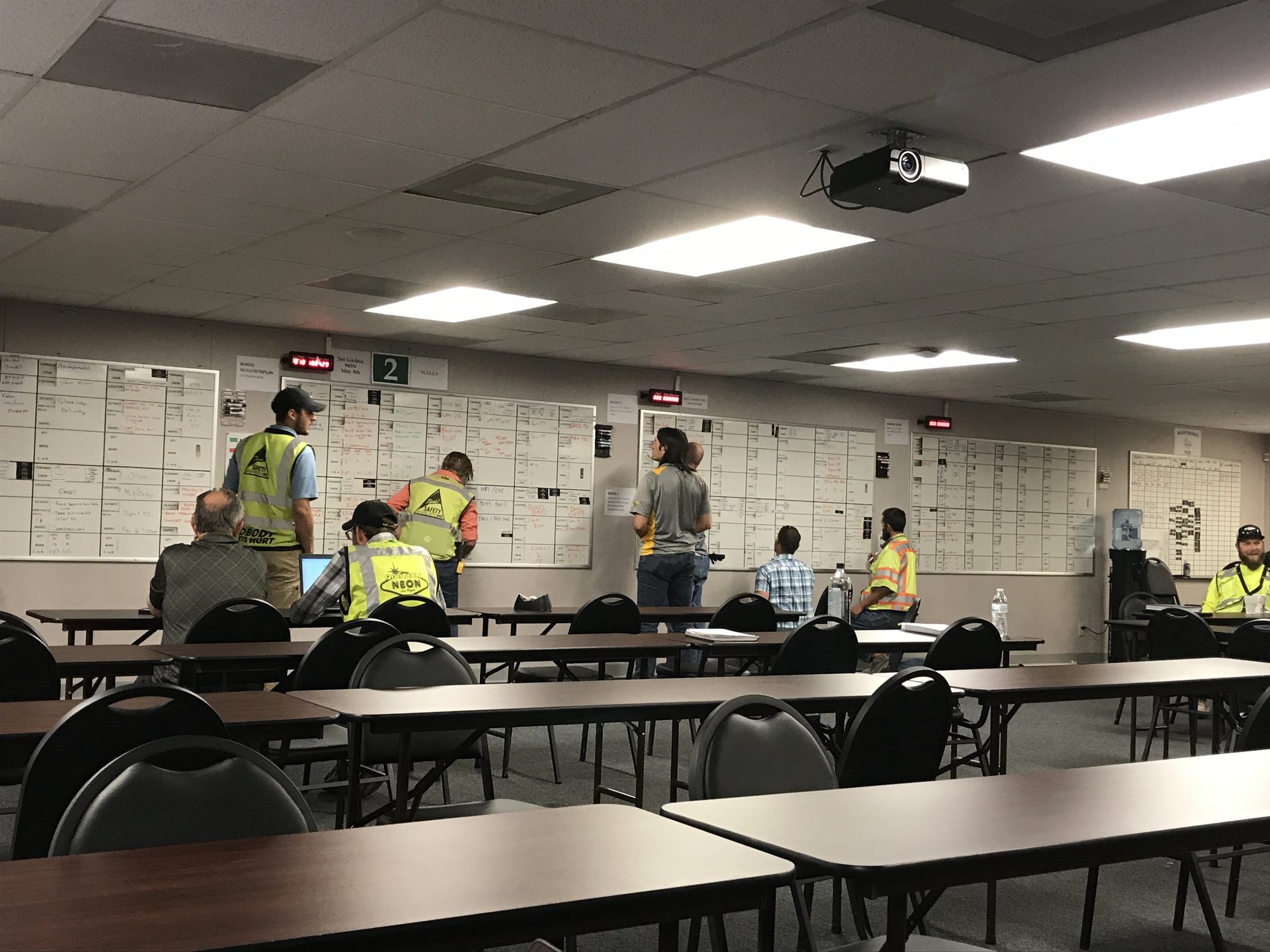 Crew members in safety vests reviewing printed schedules that cover the walls of a site office