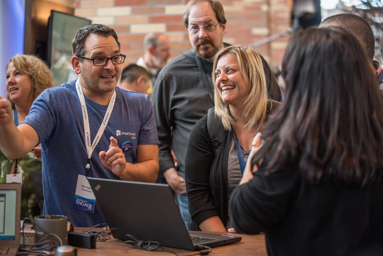 A Smartsheet staffer in a blue Smartsheet shirt demoing on a laptop to smiling attendees at the Engage conference